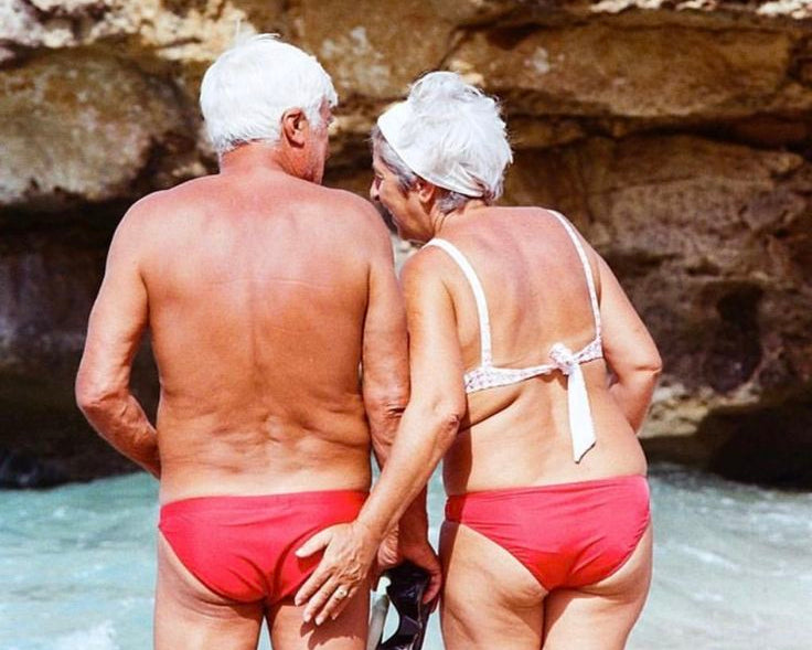 Two people in red swimwear standing close to each other by a rocky shoreline.