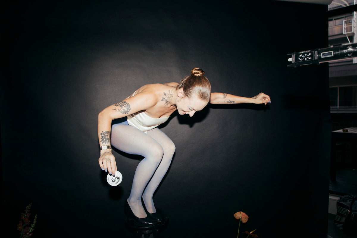 A woman crouched on a stool against a black studio backdrop, holding the tin