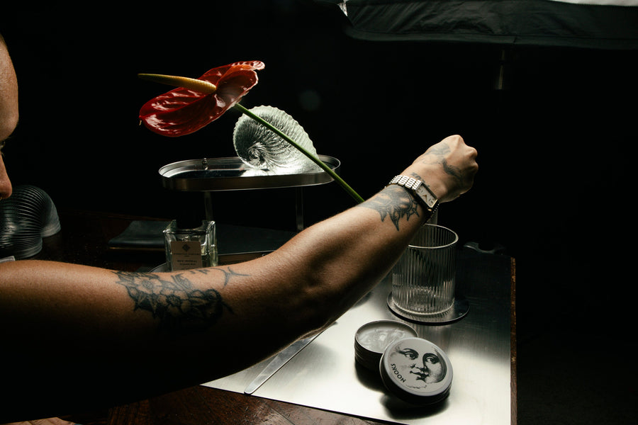 An open NOOKS tin held in a tattooed arm beside an anthurium